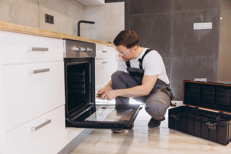 Plumber wearing safety glasses using a screwdriver to repair an electric oven in a modern kitchen, with toolbox next to him