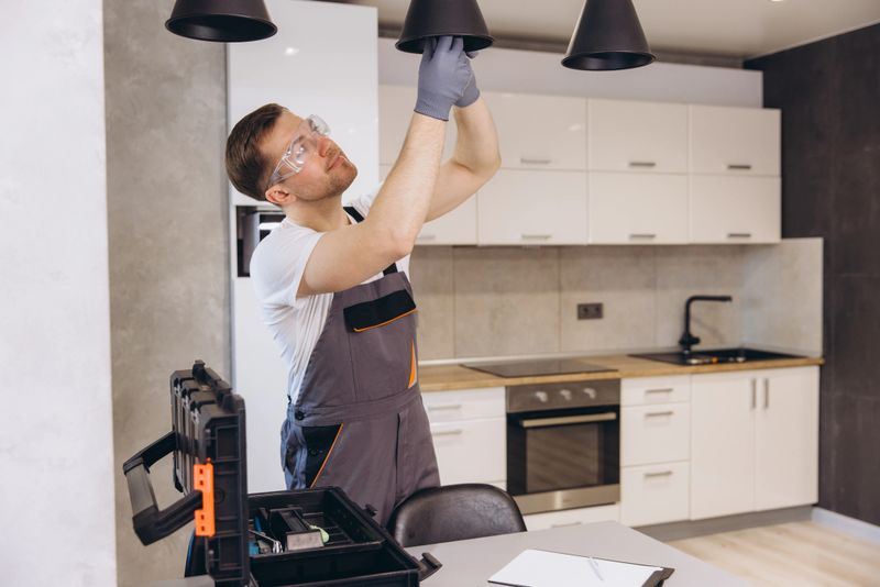Electrician wearing safety glasses and gloves installing hanging lamps in a modern kitchen, with toolbox and paperwork on the table