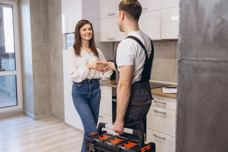 Plumber with toolbox shaking hands with homeowner in kitchen after finishing work, home repair service concept