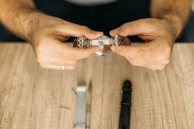 A detailed image showing hands working on a buckle of a automatic wristwatch.The scene includes tools, a wooden surface, and a focus on intricate craftsmanship, emphasizing precision and skill involved in watch repair