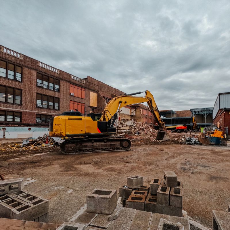 Large scale construction and demolition site with heavy equipment. Concrete bricks in the foreground of view. Backhoe with hydraulic arm is set against a partially demolished building. Cloudy blue skies above.