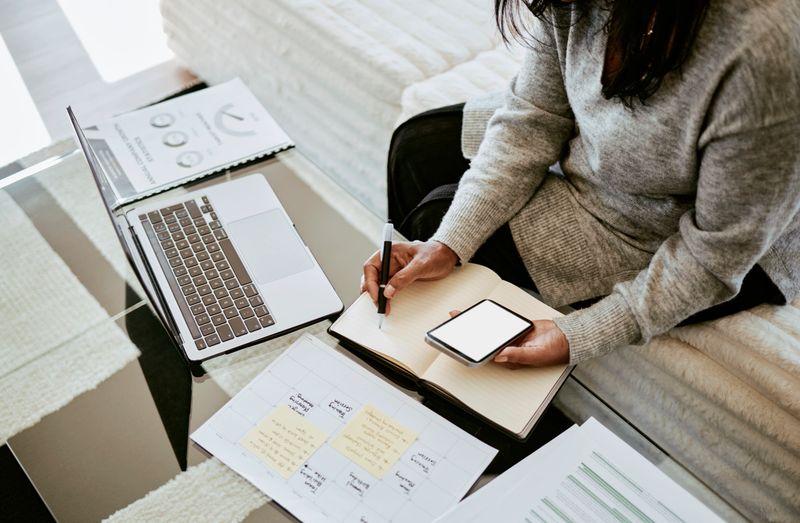 A woman writing in a notebook, using a smartphone and laptop on a glass table, surrounded by documents and a planner in a cozy workspace.