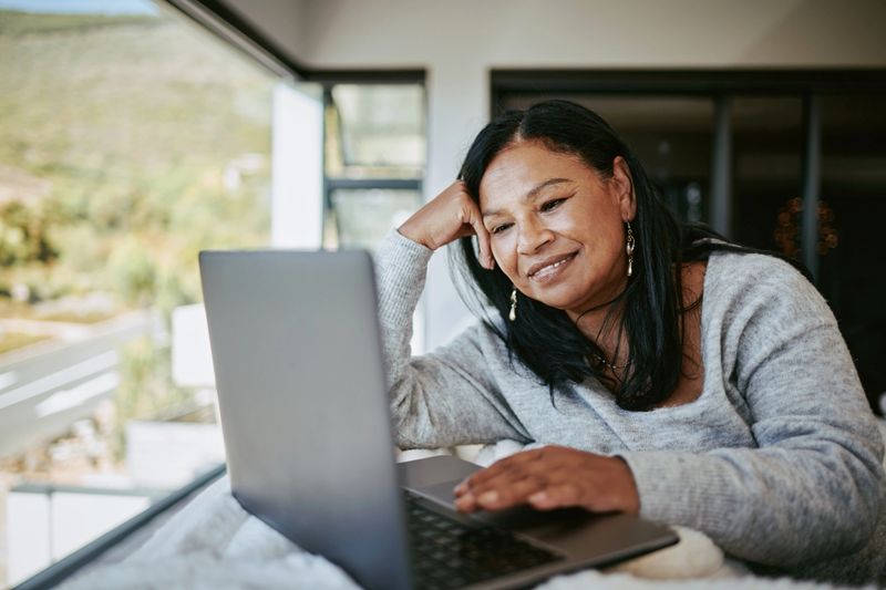 A cheerful middle-aged woman with dark hair uses a laptop while sitting on a soft white couch in a modern, well-lit room.