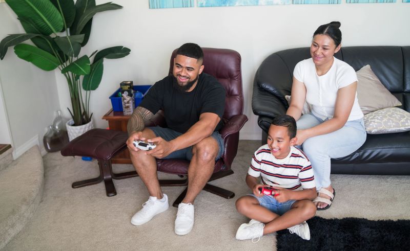 Pacific Island family enjoying video games in a cozy living room, seated on sofa and chair, smiles and concentration visible on their faces.