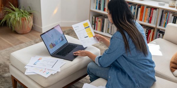 Woman reviewing bills and financial documents on a laptop in a cozy living room.