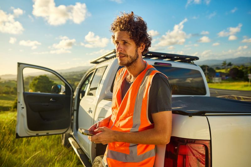Young adult engineer using smartphone while leaning on his pickup truck in rural australia, wearing high-visibility vest