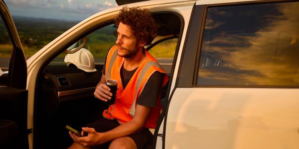 Construction worker in orange vest sitting in vehicle, holding a cup and phone at sunset.