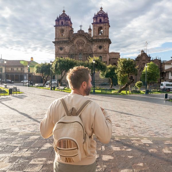 A man with a backpack stands in a sunny plaza facing a historic cathedral.