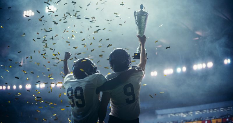 American Football Team Celebrating a Championship Victory with Golden Trophy and Falling Confetti Under Stadium Lights. Emotional Moment of Triumph for Two Diverse Footballers in White Jerseys