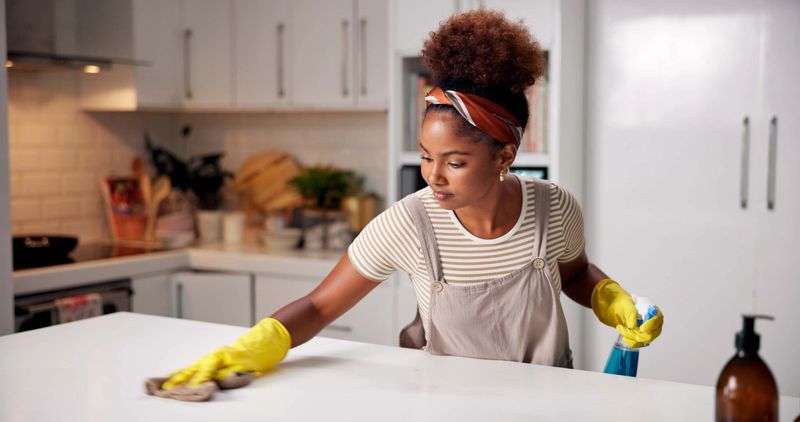 Woman, cloth and wipe on kitchen counter for hygiene with spray, gloves or chemical for shine in home. Person, cleaning and bottle for dust, dirt or bacteria with sanitation, liquid or task in house