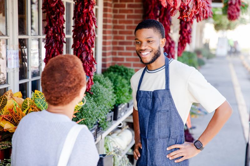 Smiling shopkeeper helps a customer outside a small market.
