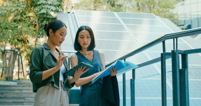 Two young Asian businesswomen review documents and digital tablet next to solar panels, collaborating on sustainable energy project in urban environment. Sustainable business outdoor concept.