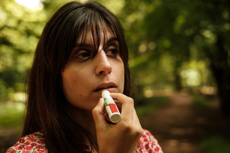 Young woman in a lush forest applying lip balm, surrounded by green trees, embracing nature's beauty. Sunlight filters through leaves, casting a serene glow on her face as she cares for her lips
