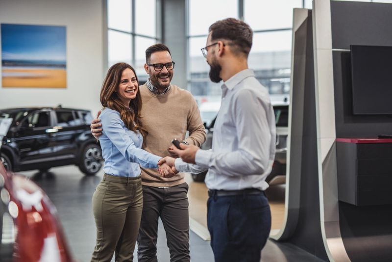 Happy couple shaking hands with a car salesman, celebrating their successful purchase of a new vehicle at the dealership, smiling and expressing excitement in the vibrant showroom