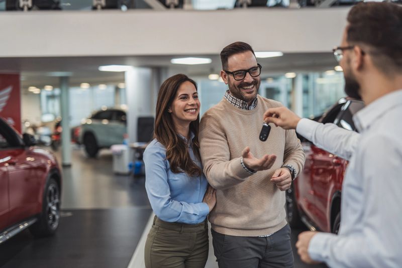 Happy couple smiling and receiving new car keys from a car salesman in a dealership, they just bought their new car and are excited to drive it home