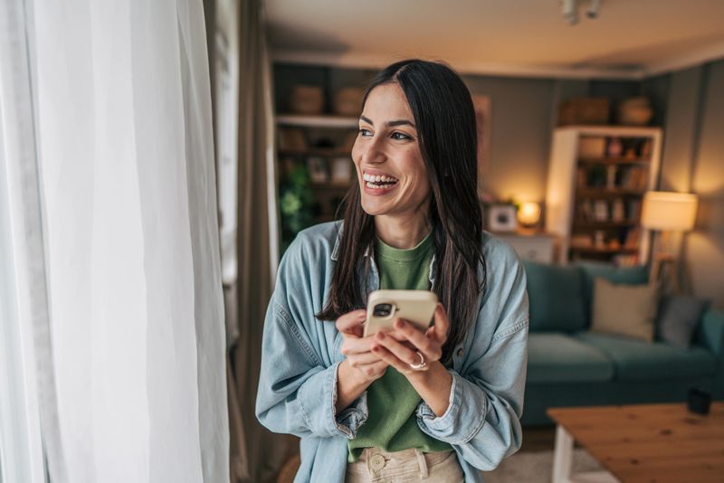 Happy young woman standing near the window in her cozy living room, smiling while using her mobile phone and gazing away, enjoying a moment of connection and relaxation