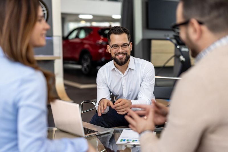 Car salesman sitting at a desk with a laptop and documents, discussing a new car purchase with a couple in a car dealership, smiling and gesturing with his hands
