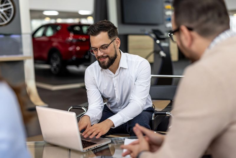 Car salesman using a laptop, sitting at a table and explaining something to a couple of customers, in a car dealership showroom with a new car in the background