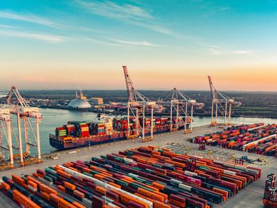 Porto comercial com guindastes e contêineres coloridos organizados no pátio, além de um navio carguei