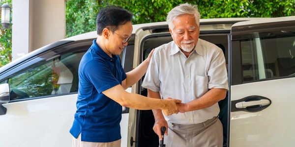 Younger man assisting elderly man with a cane while exiting a vehicle.