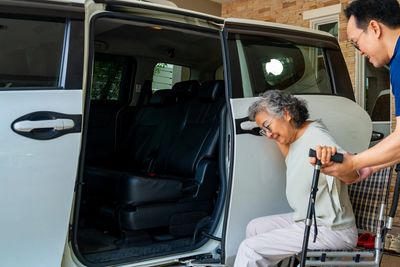 A man helps an elderly woman from a wheelchair into a car.