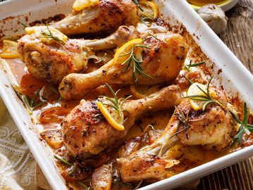 Baked chicken drumsticks garnished with lemon slices and rosemary in a white baking dish.