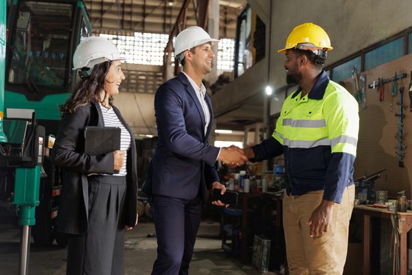 Three professionals in hard hats shaking hands in an industrial setting.