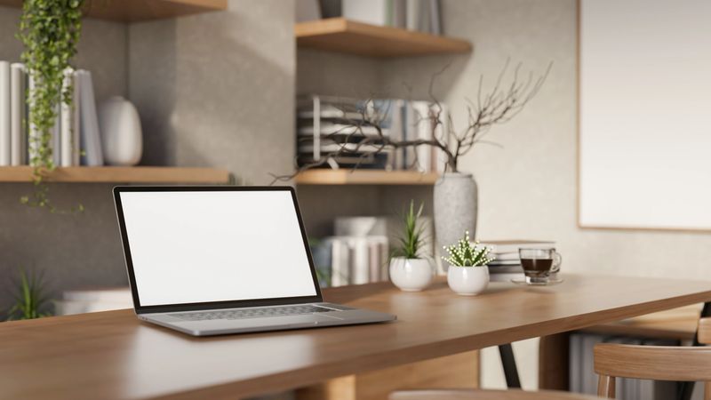 The white screen laptop and a coffee cup on a long wooden table with decors in the bright room. Study corner set up, 3D Rendering, 3D Illustration.