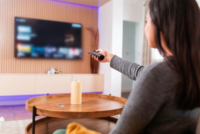 A woman sits comfortably in her living room, controlling the TV with a remote. The scene captures a relaxed, cozy atmosphere with home interior details including a stylish coffee table and warm lighting.