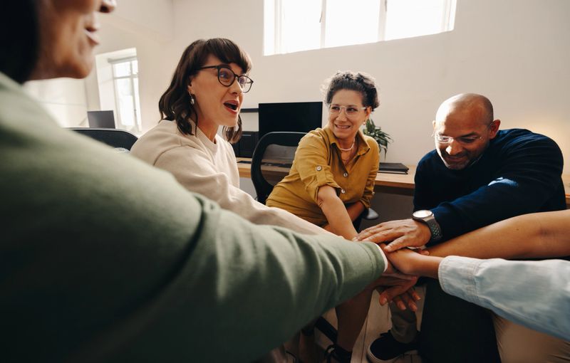 A team engaged in a collaborative discussion, showing unity and enthusiasm by joining hands together. The image reflects a professional work environment focused on teamwork and achieving shared goals.