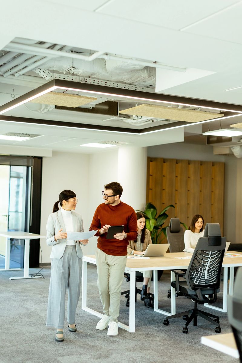 Two colleagues engage in a dynamic discussion while walking through a contemporary office filled with creativity. Others focus on their tasks, showcasing teamwork.