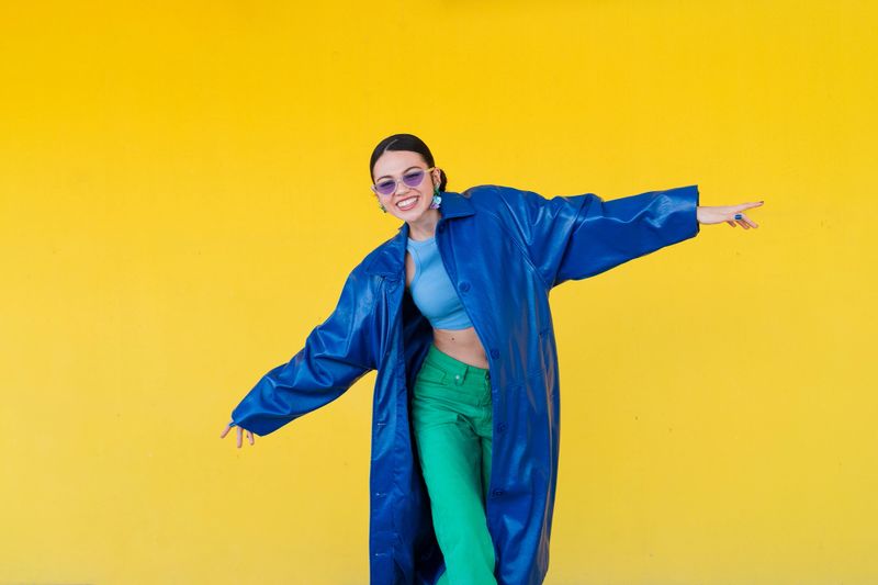 Fashionable young woman wearing a blue coat and sunglasses strikes a pose on a vibrant yellow background, exuding confidence and style
