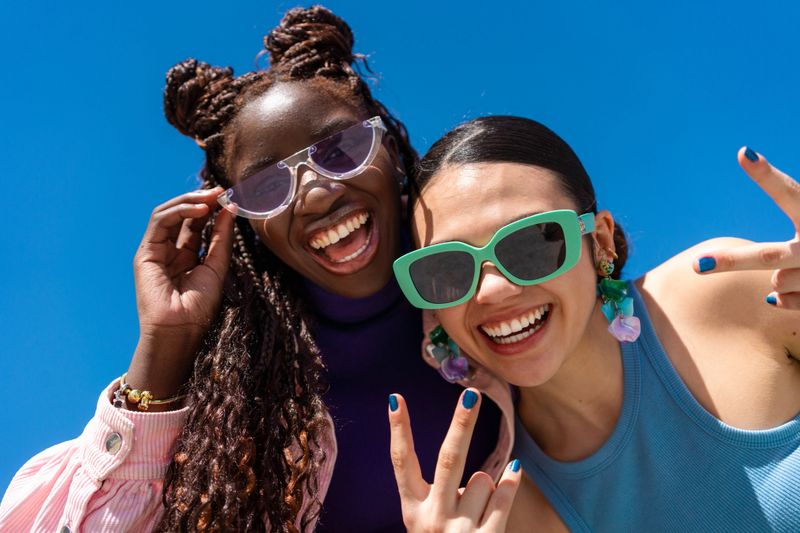 Cheerful young women wearing sunglasses, flashing peace signs while standing together under bright blue sky backdrop