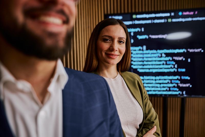 Two software developers, a man and a woman, smiling and looking confidently at the camera while standing in front of code displayed on a monitor in a modern office environment