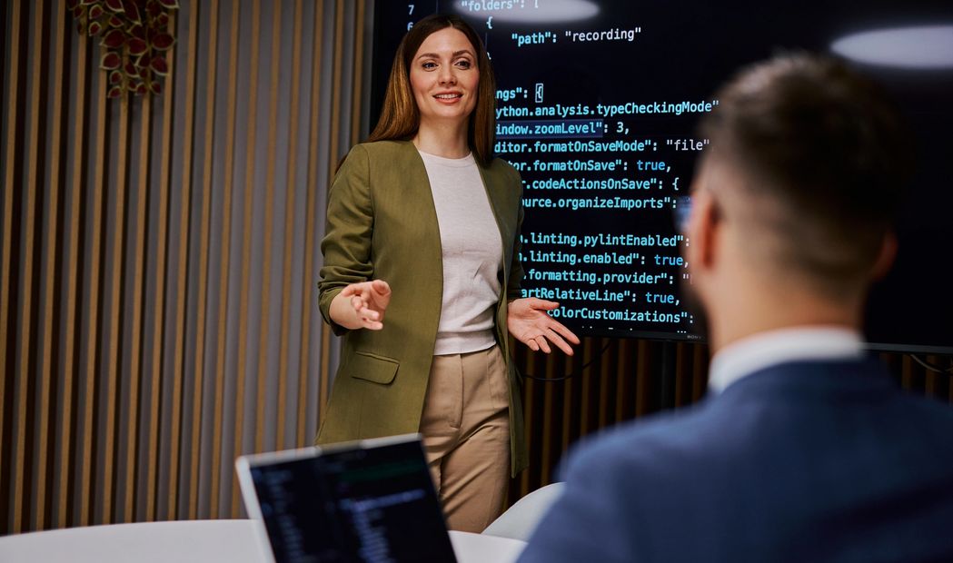 A woman presents coding details on a screen to a colleague in a modern office.