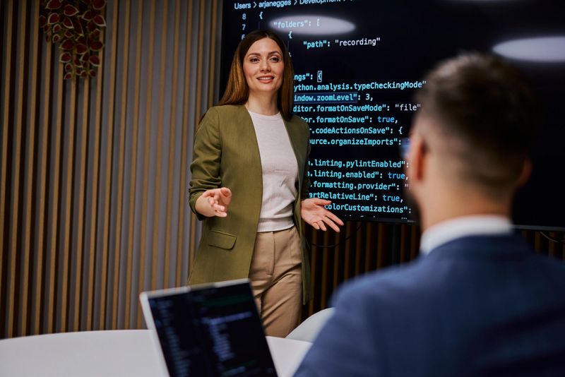 Female software developer is presenting code on a monitor to her colleague during a business meeting, discussing programming and development strategies