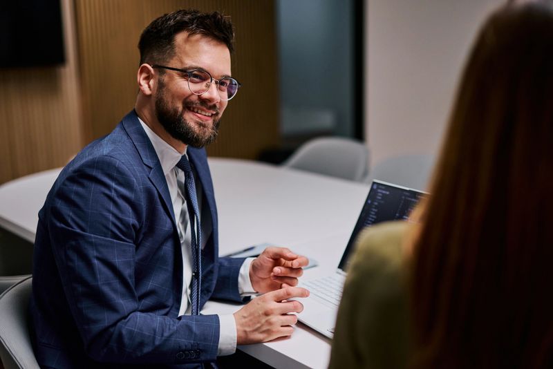 Businessman in a suit and tie sitting at a table, smiling and engaging in conversation with a female colleague during a productive business meeting in a modern office setting