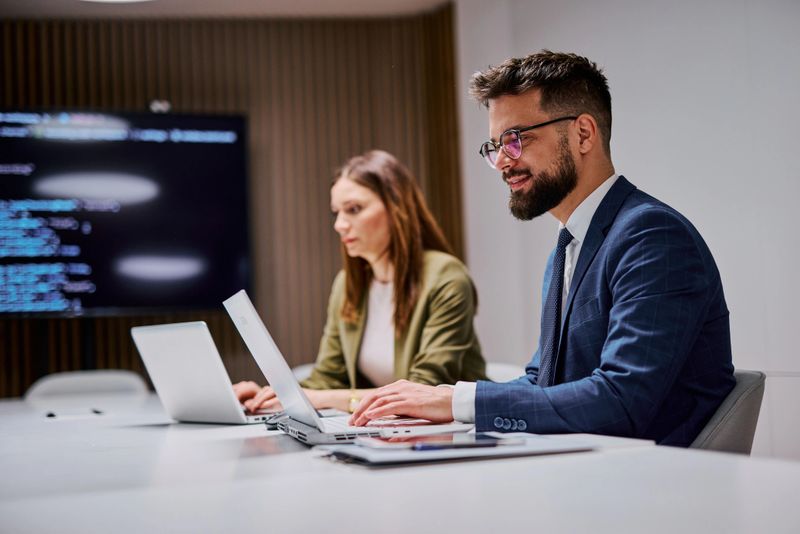 Businessman and businesswoman are working on their laptops in a modern office, collaborating on a project with code displayed on a screen in the background