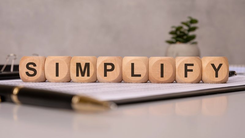 The word SIMPLIFY is spelled with wooden cubes on office papers, with pens and a potted plant in the background, symbolizing efficiency, minimalism, productivity, clarity, and organization