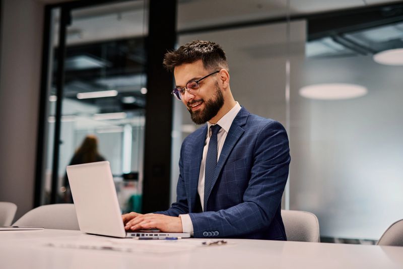 Young businessman wearing glasses and a suit, sitting at a sleek desk in a modern office, smiling while working on a laptop and engaging with digital tasks and projects