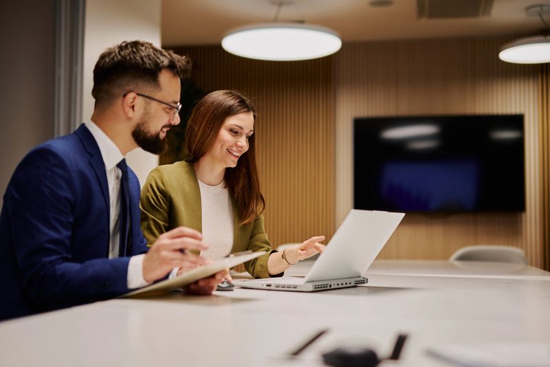 Two young professionals collaborating on a project, engaging with a laptop and taking notes, surrounded by a bright, modern office space that fosters creativity and teamwork