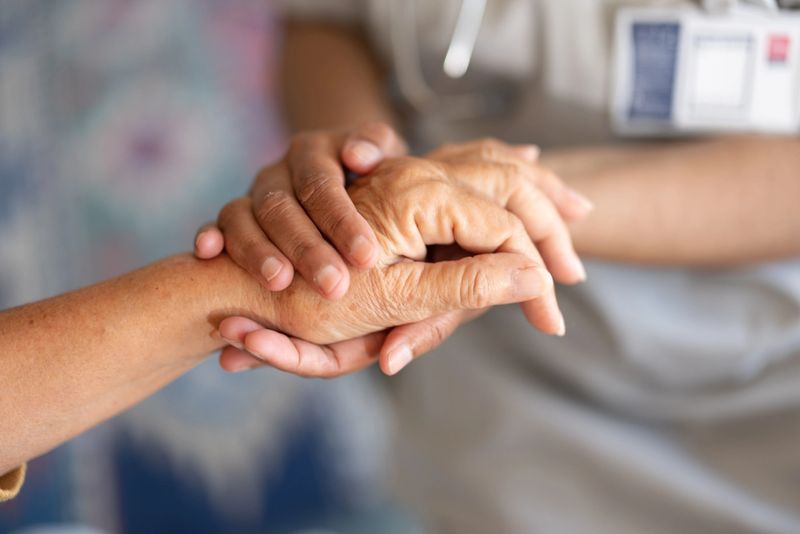A close-up of a senior woman’s hands being gently held by a caregiver or nurse.  The warm home setting with natural light symbolizes care, comfort, support, and emotional connection.
