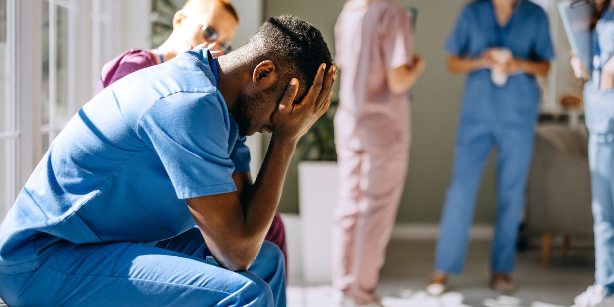 A stressed healthcare worker sits with head in hands, while colleagues chat in the background.