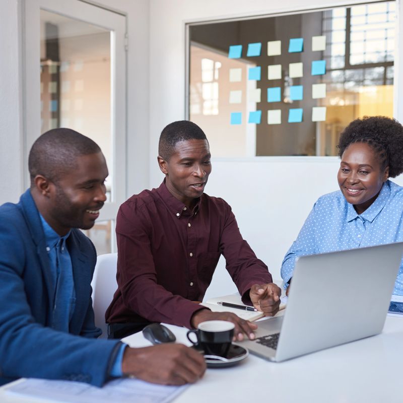 Three casually dressed African colleagues talking together while sitting at a desk in a modern office working on a laptop