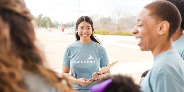 Smiling volunteer woman with clipboard outdoors, talking to a group.