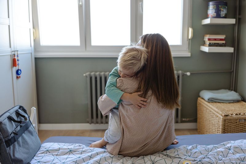 A mother embraces her child while seated on a bed in a peaceful bedroom environment. The atmosphere evokes feelings of love, care, and warmth, capturing a tender and emotional moment.