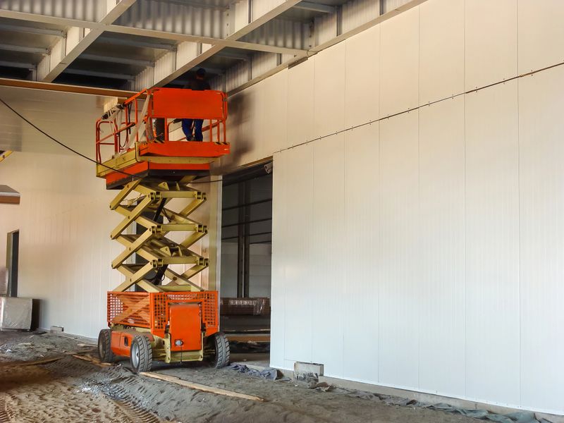 Workers are seen on a scissor lift installing insulated metal panels on the interior of a building under construction.