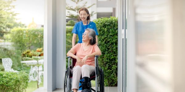 A nurse aid is pushing a wheelchair that is carrying an elderly person, and both are smiling. The lo