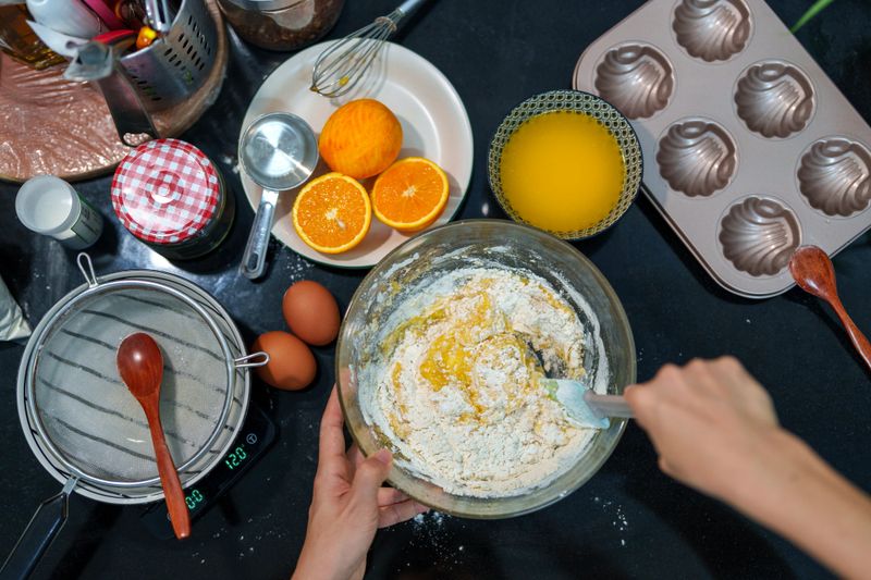 A person stirs a fresh orange-infused batter in a glass bowl, preparing for baking. Halved oranges, eggs, and a muffin tray sit on the kitchen counter, creating a cozy homemade baking scene.