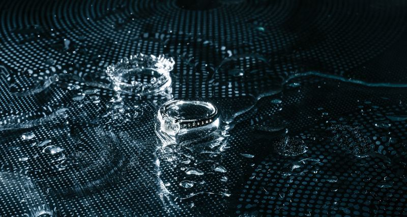 Close-up of a diamond ring surrounded by water droplets on a textured surface, highlighting intricate details and the interplay of light on the jewelry and background